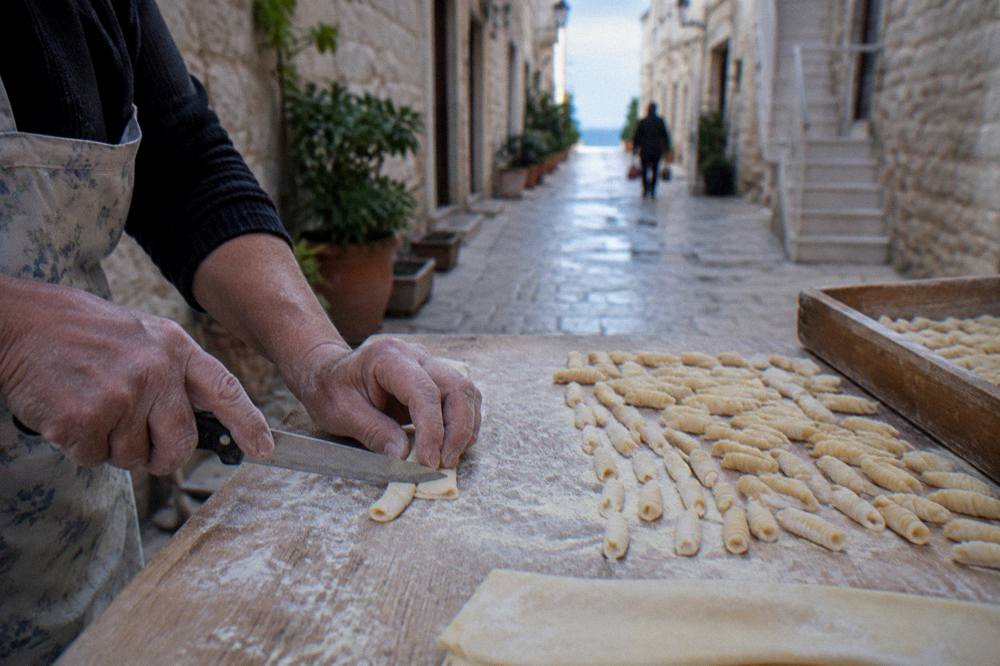 Floury hands drawing a small piece of dough across a board with a knife beside rows of finished pasta