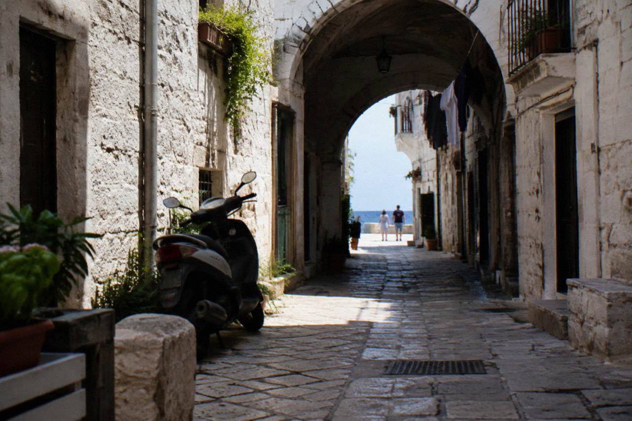 Straight old town lane between whitewashed walls ending in a bright slit of open sea with a parked scooter