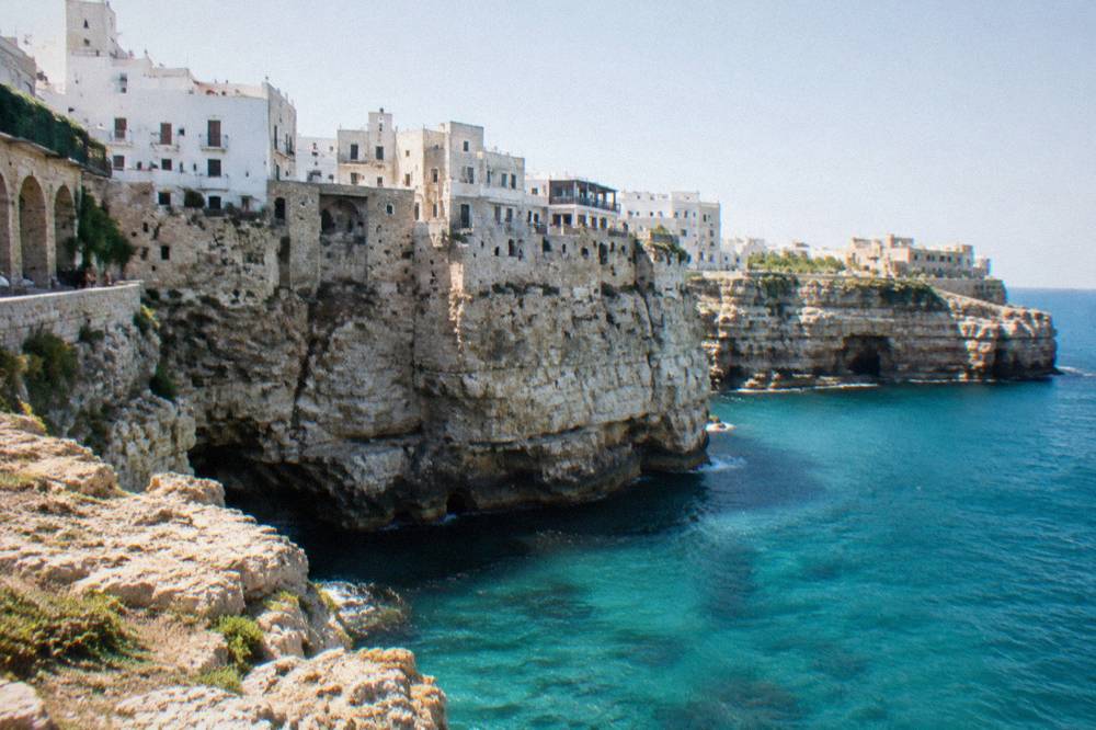 White town perched on the edge of tall pale limestone cliffs above a turquoise sea seen from a rocky viewpoint