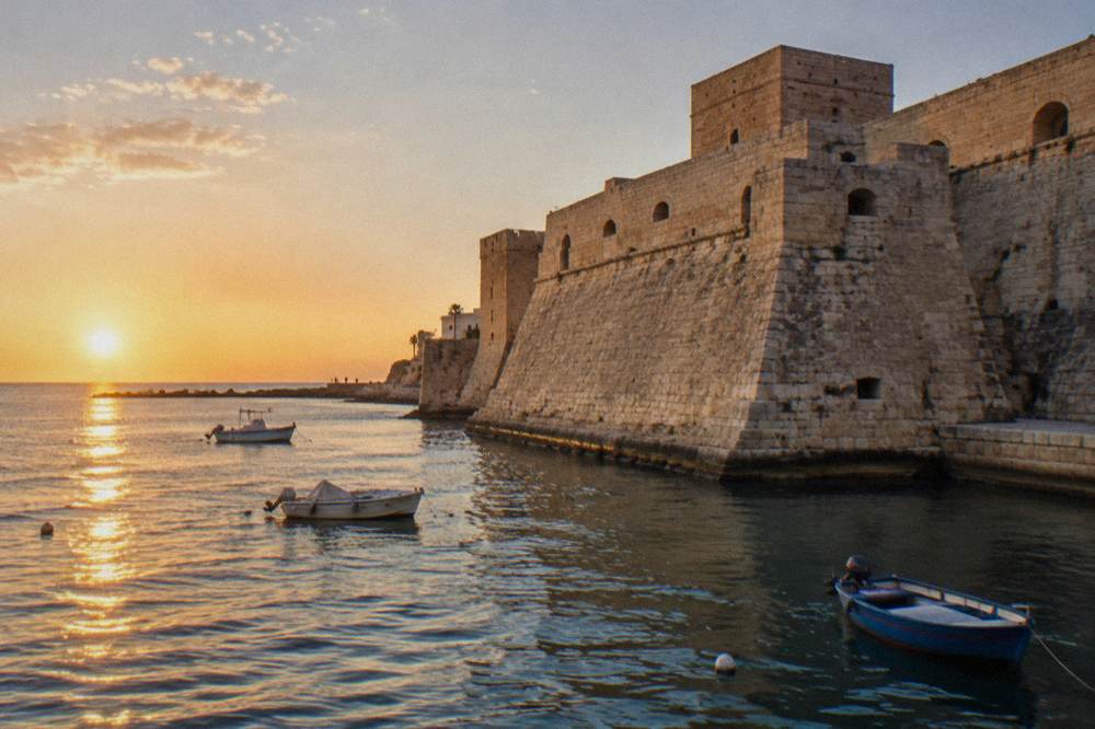 Seaward wall of a pale stone castle beside harbour water with a few small boats in afternoon sun