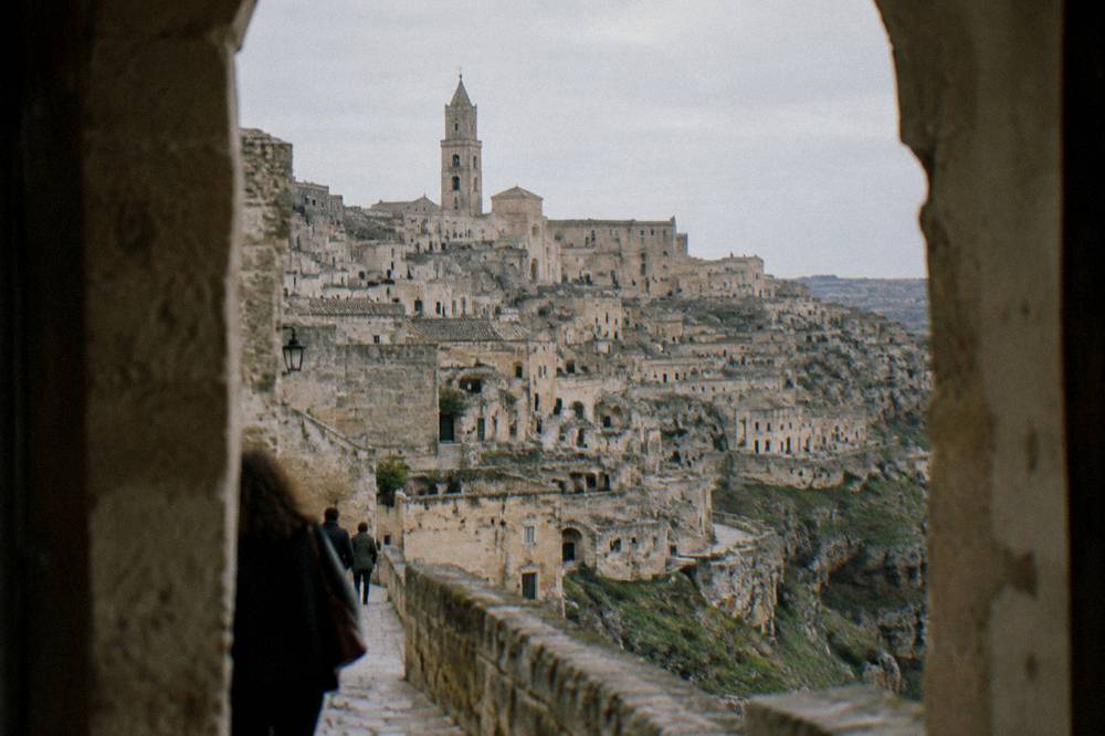 Tiers of honey-coloured cave dwellings stacked on the side of a deep ravine below a cathedral bell tower