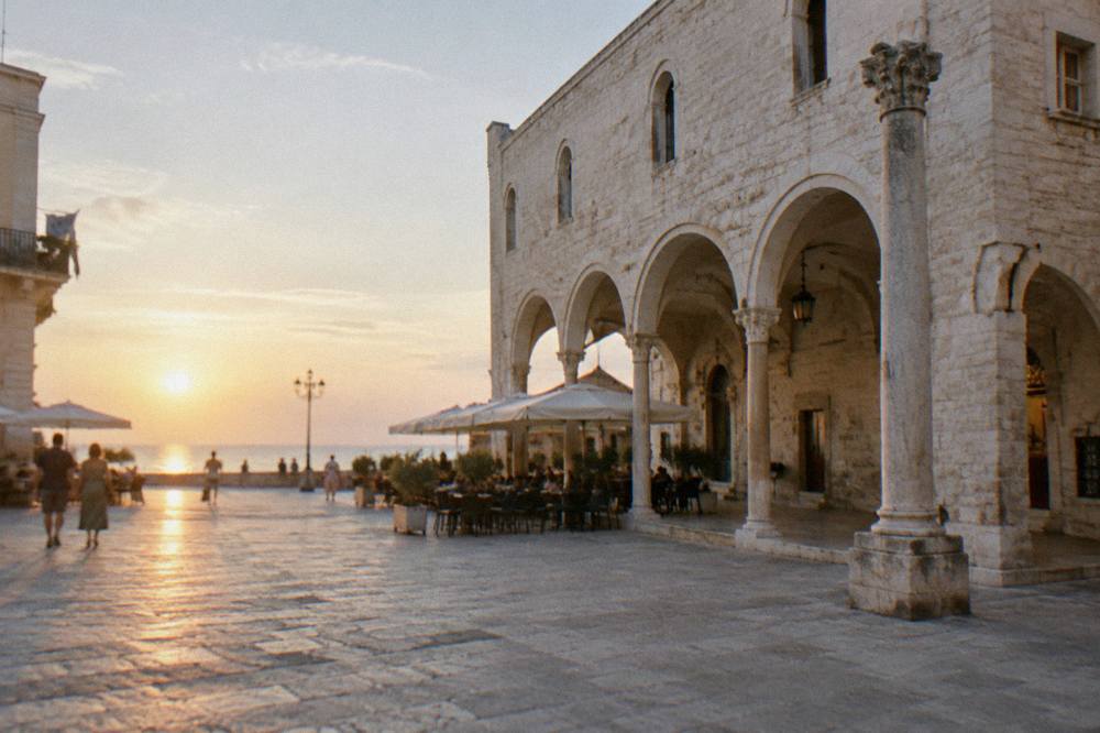 Old town square with a medieval arcaded loggia, a slender stone column and cafe tables under parasols in soft morning light