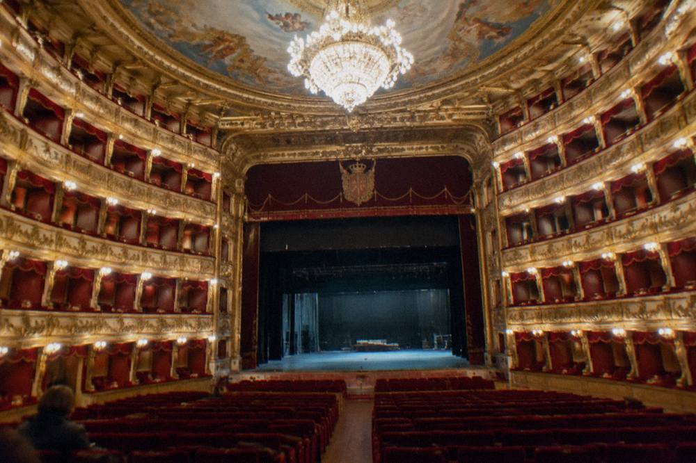 Empty horseshoe-shaped theatre with tiers of red and gold boxes, a large chandelier and a painted ceiling