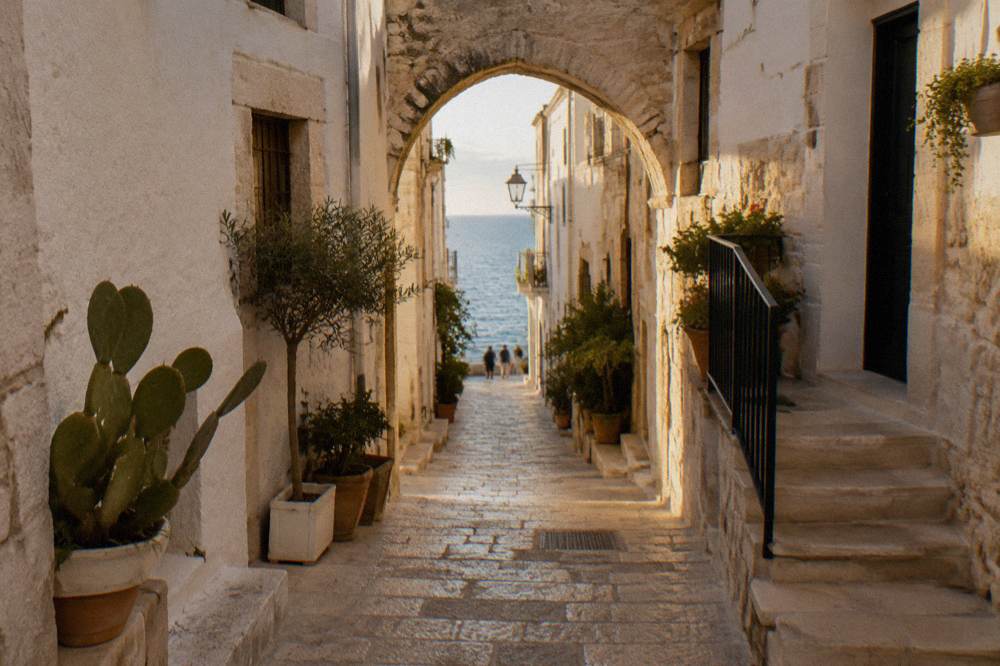 Narrow whitewashed cliff-top old town lane with a stone arch, potted plants and a glimpse of blue sea