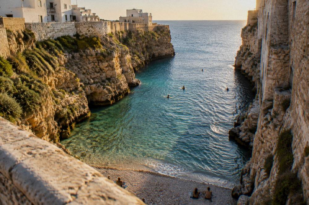 View down from a stone bridge into a narrow cove of clear turquoise water and white pebbles