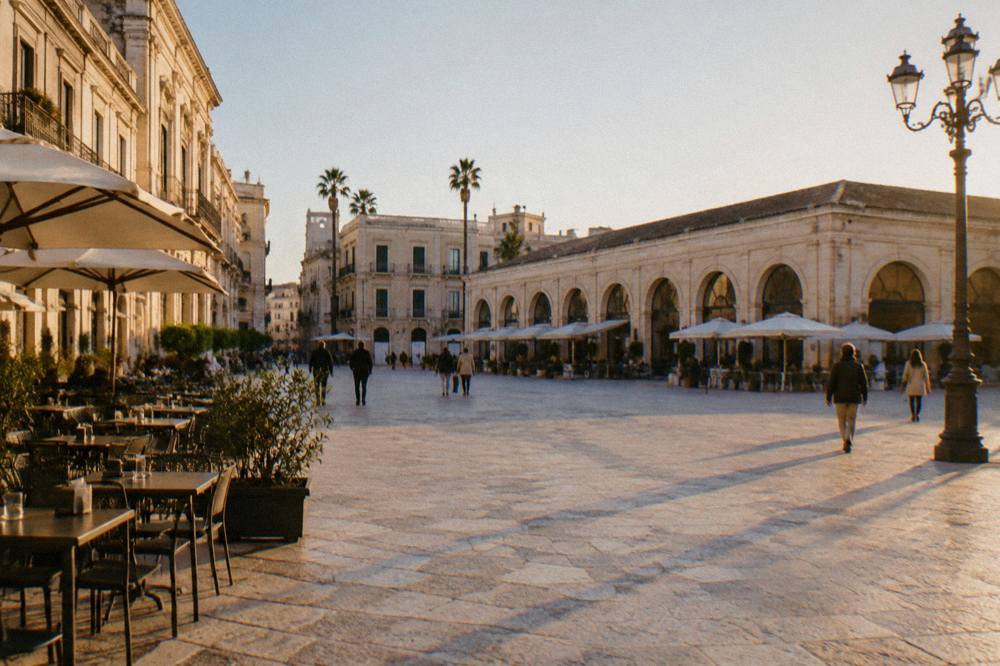 Broad open piazza beside a long low market hall with arched openings and outdoor cafe tables in bright morning